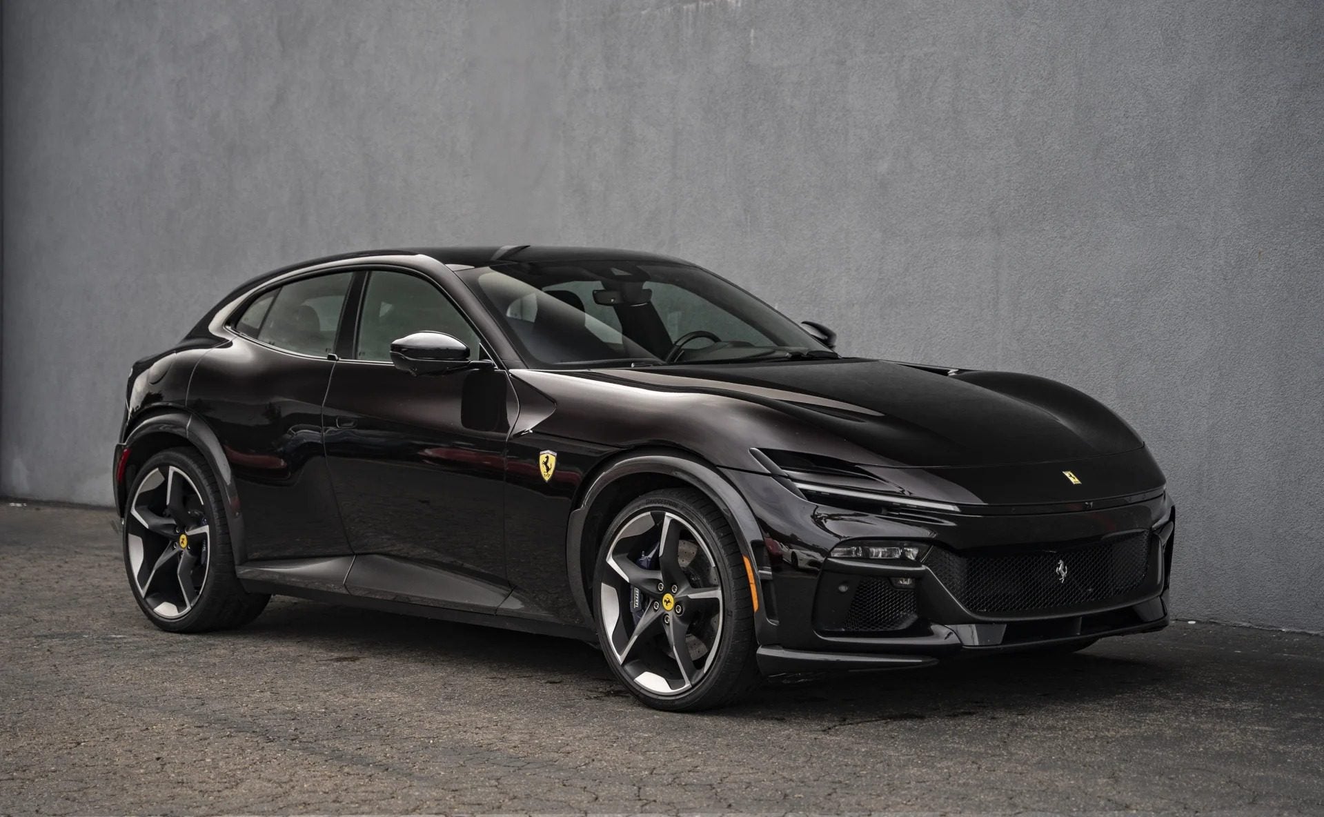 A black Ferrari Purosangue SUV is parked next to a gray concrete wall on a paved surface, viewed from the front left side.