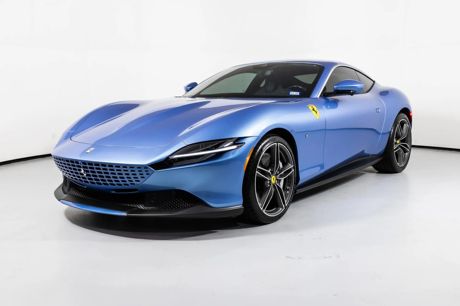 A blue Ferrari Roma, embodying La Nuova Dolce Vita, is parked in a white studio and viewed from the front left angle.