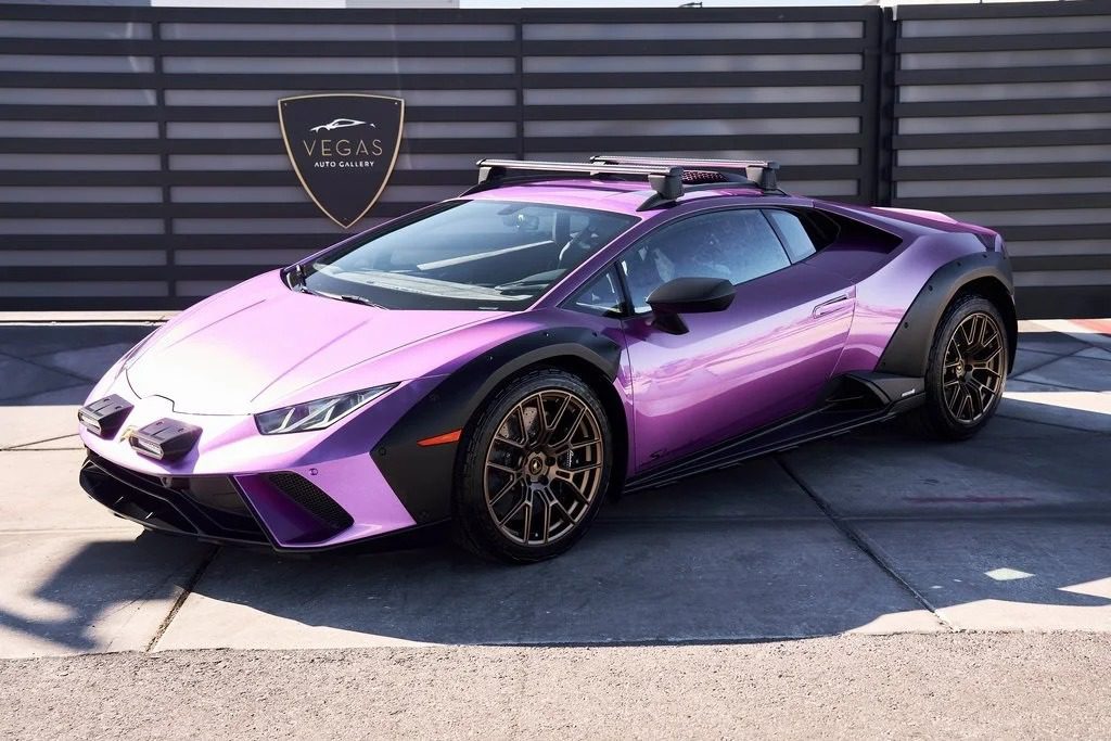 A metallic purple Lamborghini Huracán V10 supercar with roof racks and off-road modifications is parked in front of a sign that reads “Vegas Auto Gallery,” where this stunning vehicle is for sale.