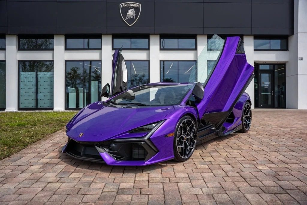 A purple Lamborghini Revuelto with its scissor doors open is parked on a brick driveway in front of a dealership, showcasing one of the best Lamborghini supercars for sale.