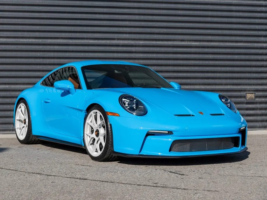 A stunning Porsche 911 S/T in striking PTS Riviera Blue, complete with white wheels, is parked gracefully on a concrete surface, set against a dark, ridged metallic wall. This eye-catching sports car is available for sale to the discerning enthusiast.