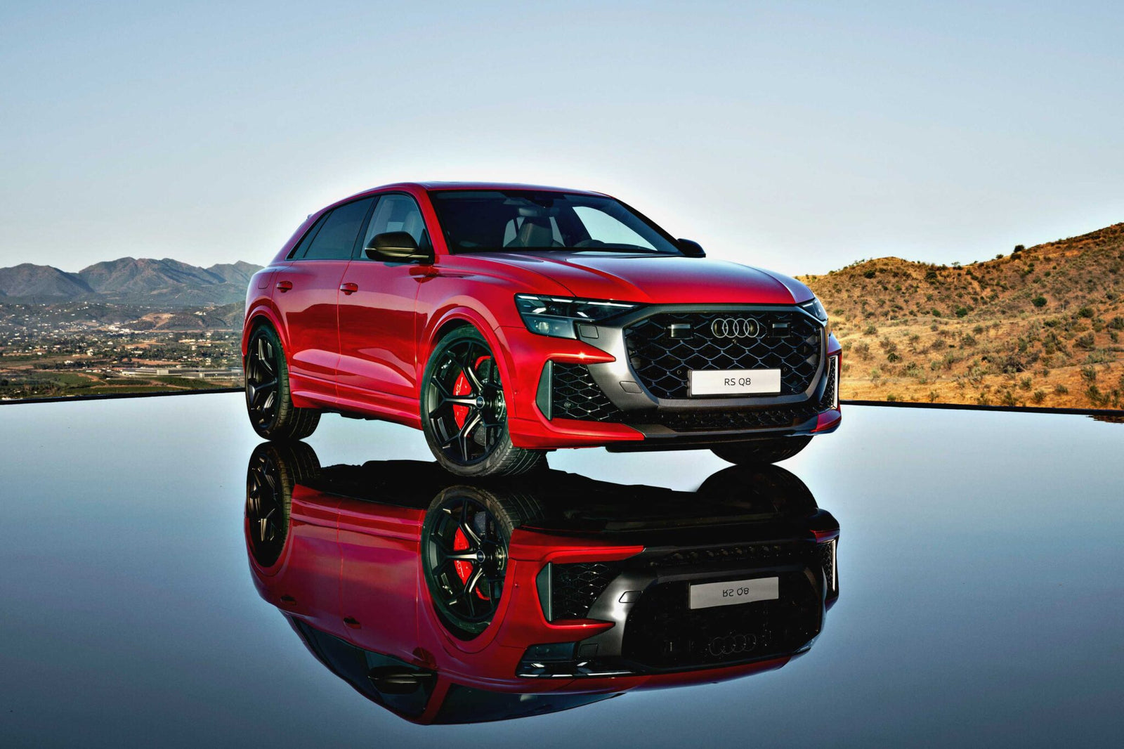 Review: 2025 Audi RS Q8 Performance—A striking red SUV parked on a reflective surface, set against a hilly landscape and clear sky.