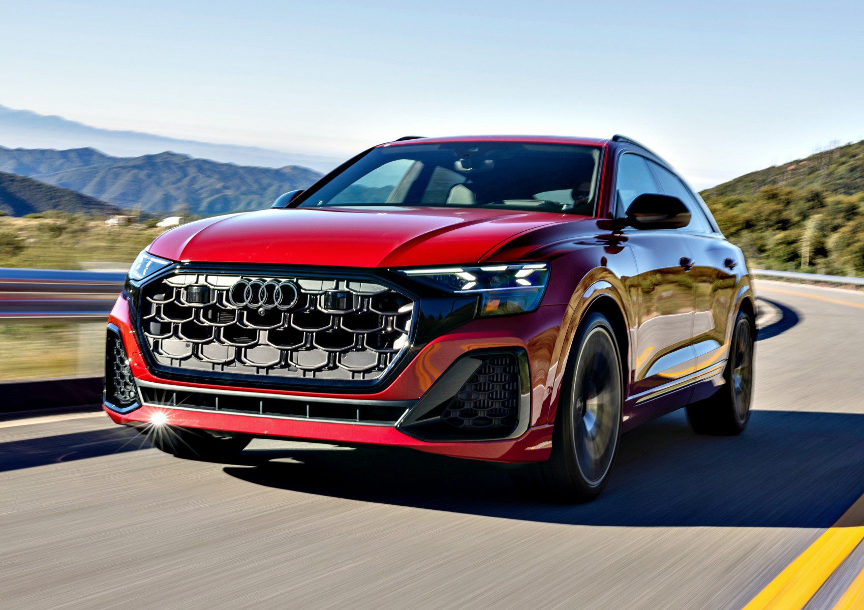 A red 2025 Audi SQ8 SUV drives on a winding road with mountains and trees in the background, showcasing its impressive style under a clear sky.