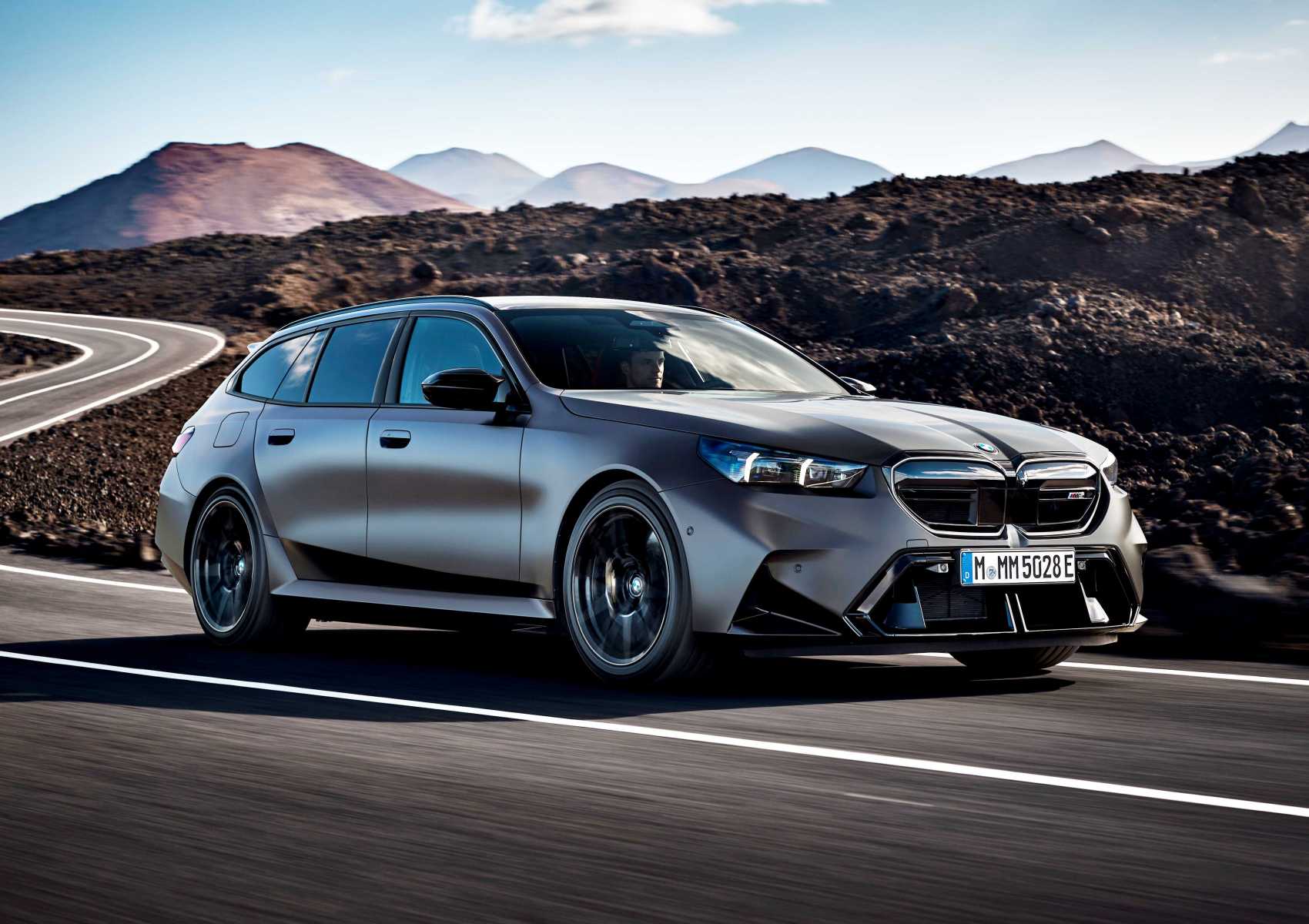A matte gray BMW station wagon drives on a winding road through a mountainous, rocky landscape under a clear sky—Review: 2025 BMW M5 Touring weighs in as a great allrounder for any adventure.