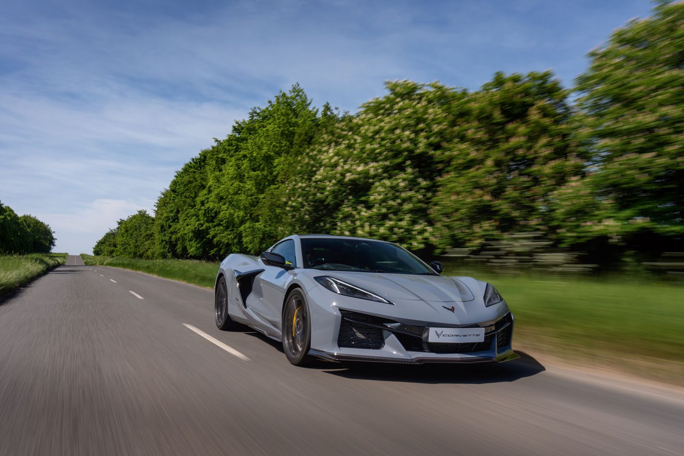 A silver sports car drives down an empty rural road lined with green trees under a blue sky.