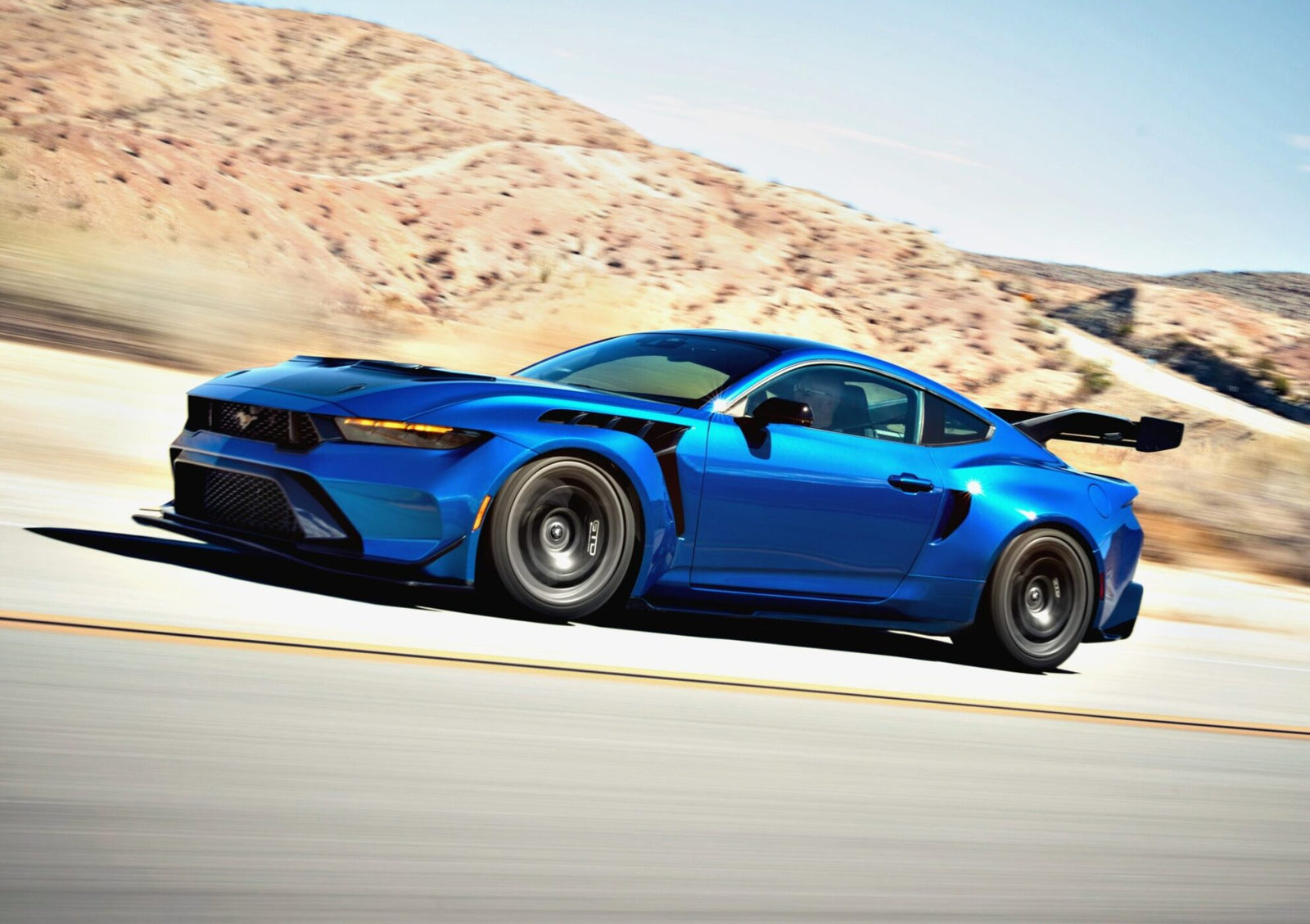 First Drive: Six-figure Ford Mustang GTD Boasts Peak Pony Muscle as a blue sports car with a large rear spoiler powers down the road, mountains rising in the background.