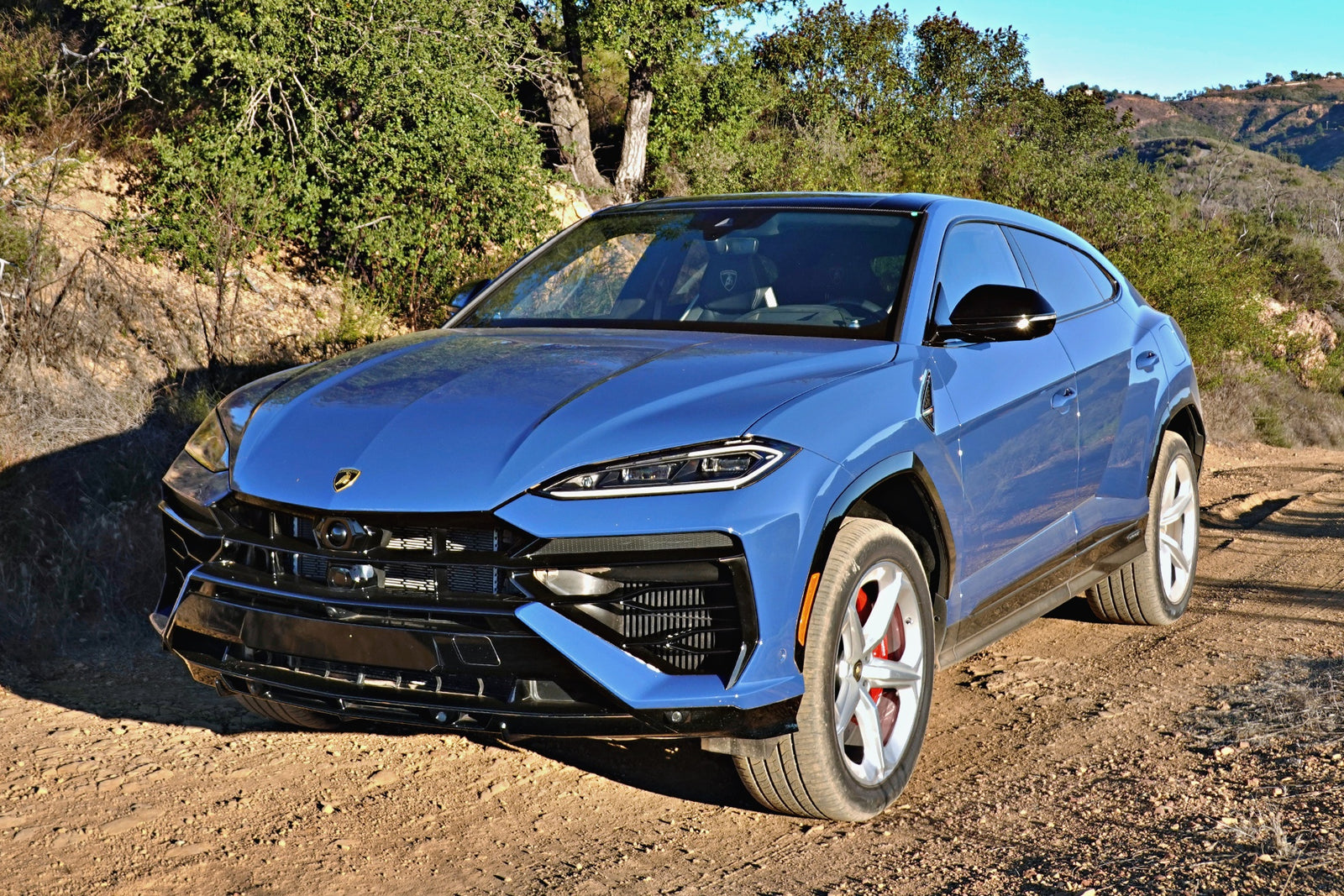 A blue 2025 Lamborghini Urus SE, the heavy hitter delivering plug-in hybrid punch, is parked on a dirt road with red brake calipers, framed by trees and hills in the background.