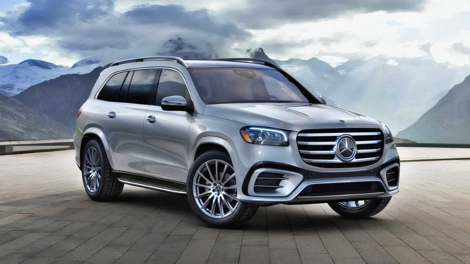 A silver Mercedes-Benz SUV, featured in the Review: 2025 Mercedes-Benz GLS 580 4Matic, is parked on a stone pavement with mountains and cloudy skies in the background.