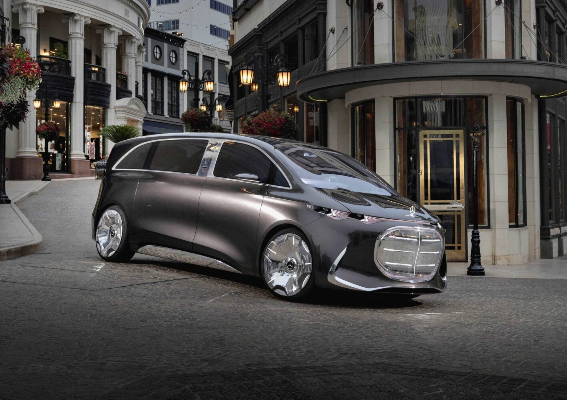 A futuristic silver Mercedes-Benz Vision V concept minivan with large wheels is parked on a cobblestone street in an upscale urban shopping area.