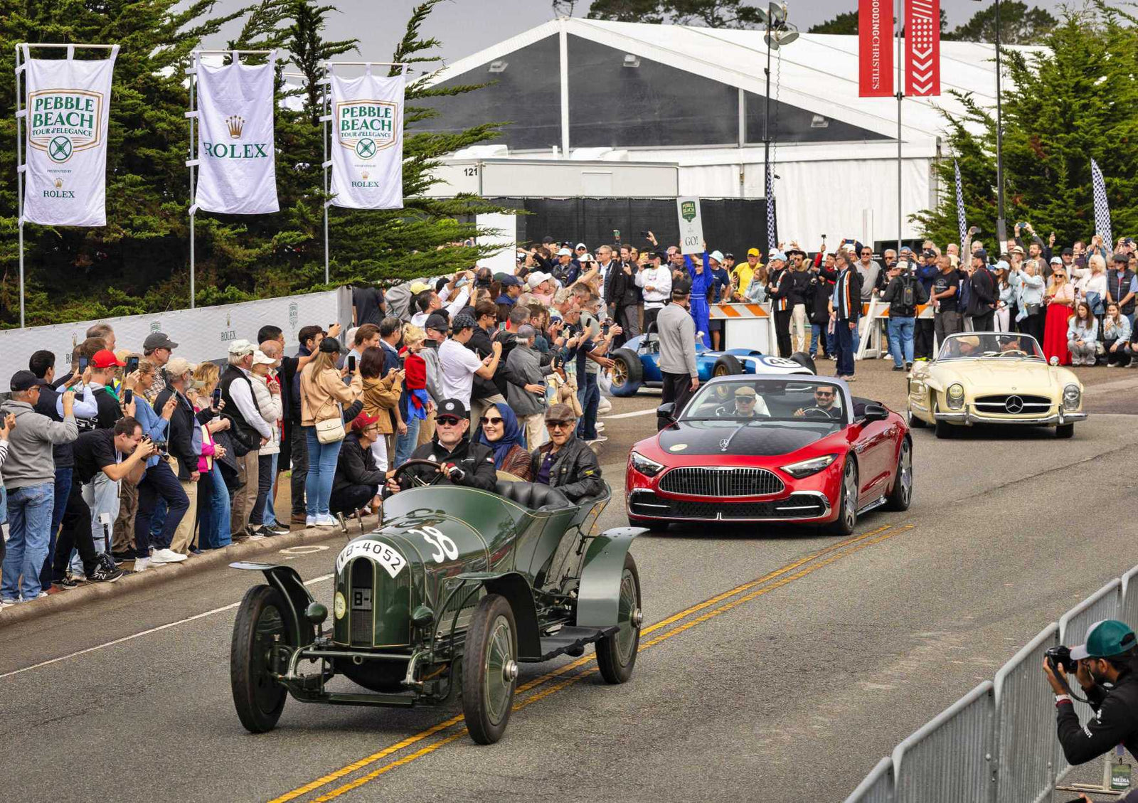 A parade of classic and modern cars, including Silver Arrows Hollywood Cars and Concept Cars: Mercedes’ Pebble Beach Highlights, drives past cheering spectators at an outdoor auto event, with banners and tents visible in the background.