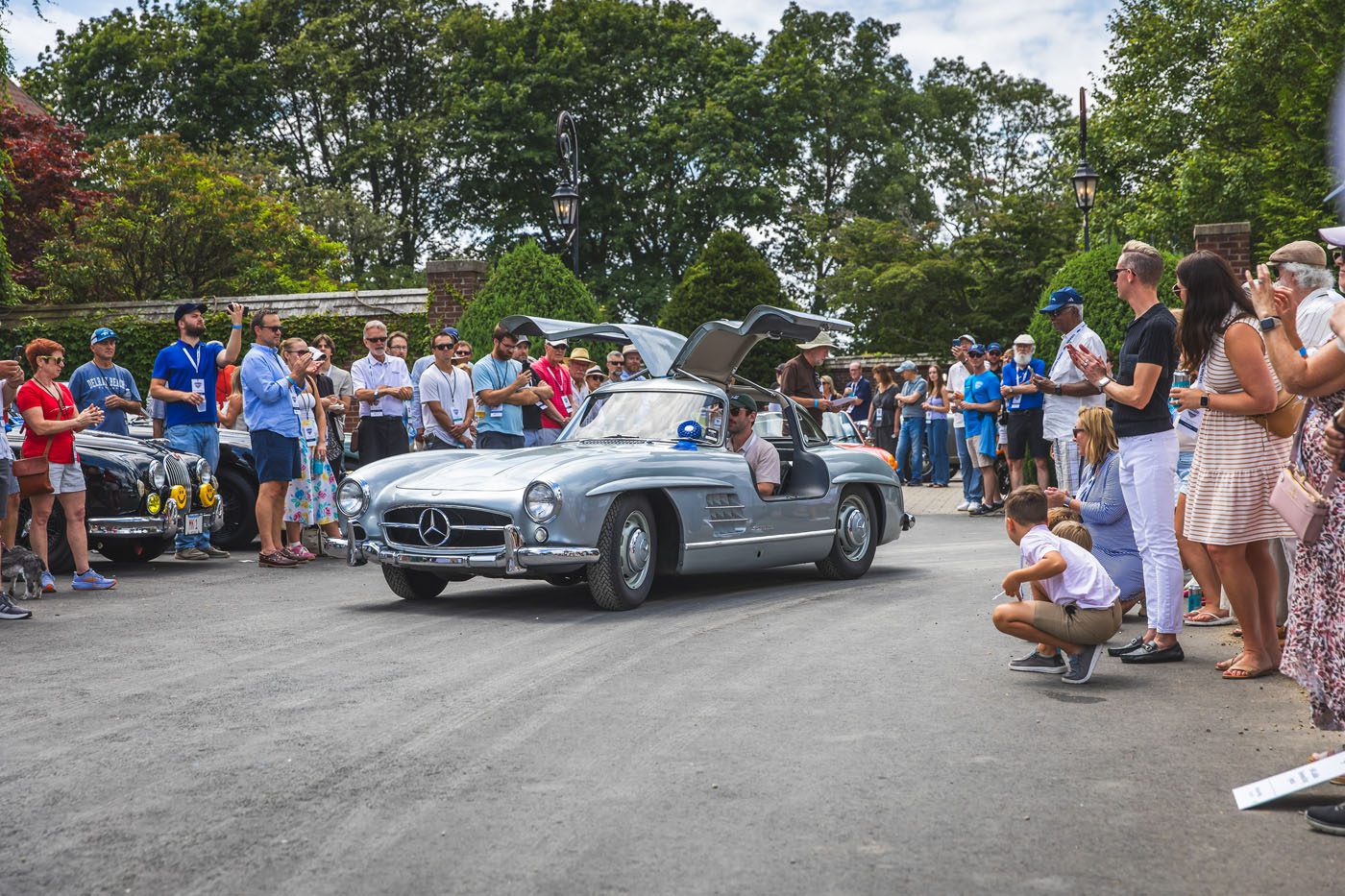 A silver vintage Mercedes-Benz with gullwing doors open drives on a road, surrounded by a crowd of people at the Misselwood Concours d’Elegance, celebrating its 15th Anniversary with record attendance and eager photographers.