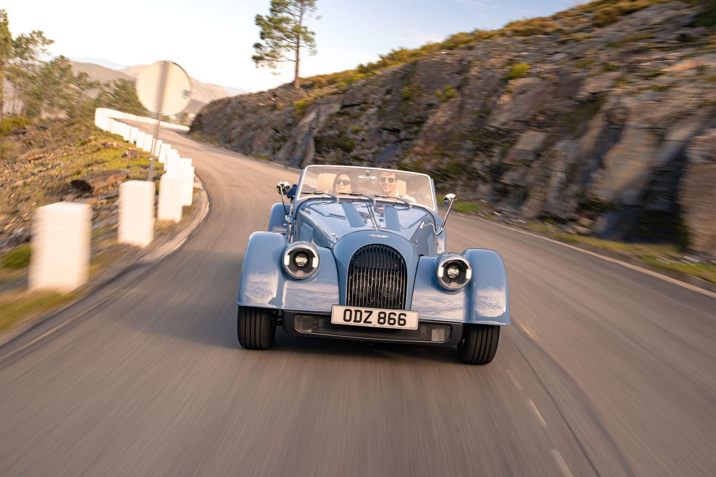 A light blue Morgan Plus Four vintage-style convertible car drives along a winding mountain road with rocky hills in the background, celebrating Morgan’s 75th Anniversary.