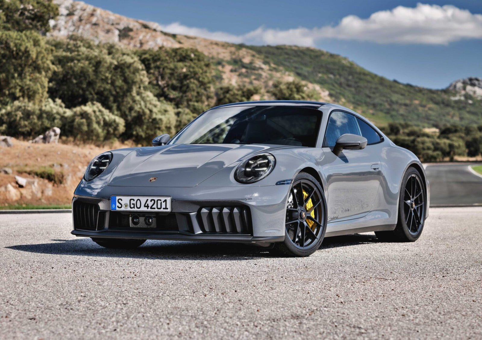 A grey Porsche sports car, featured in the Review: 2025 Porsche 911 Carrera GTS T-Hybrid Electrifies Potent Performance, is parked on a paved road with green hills and a partly cloudy sky in the background.