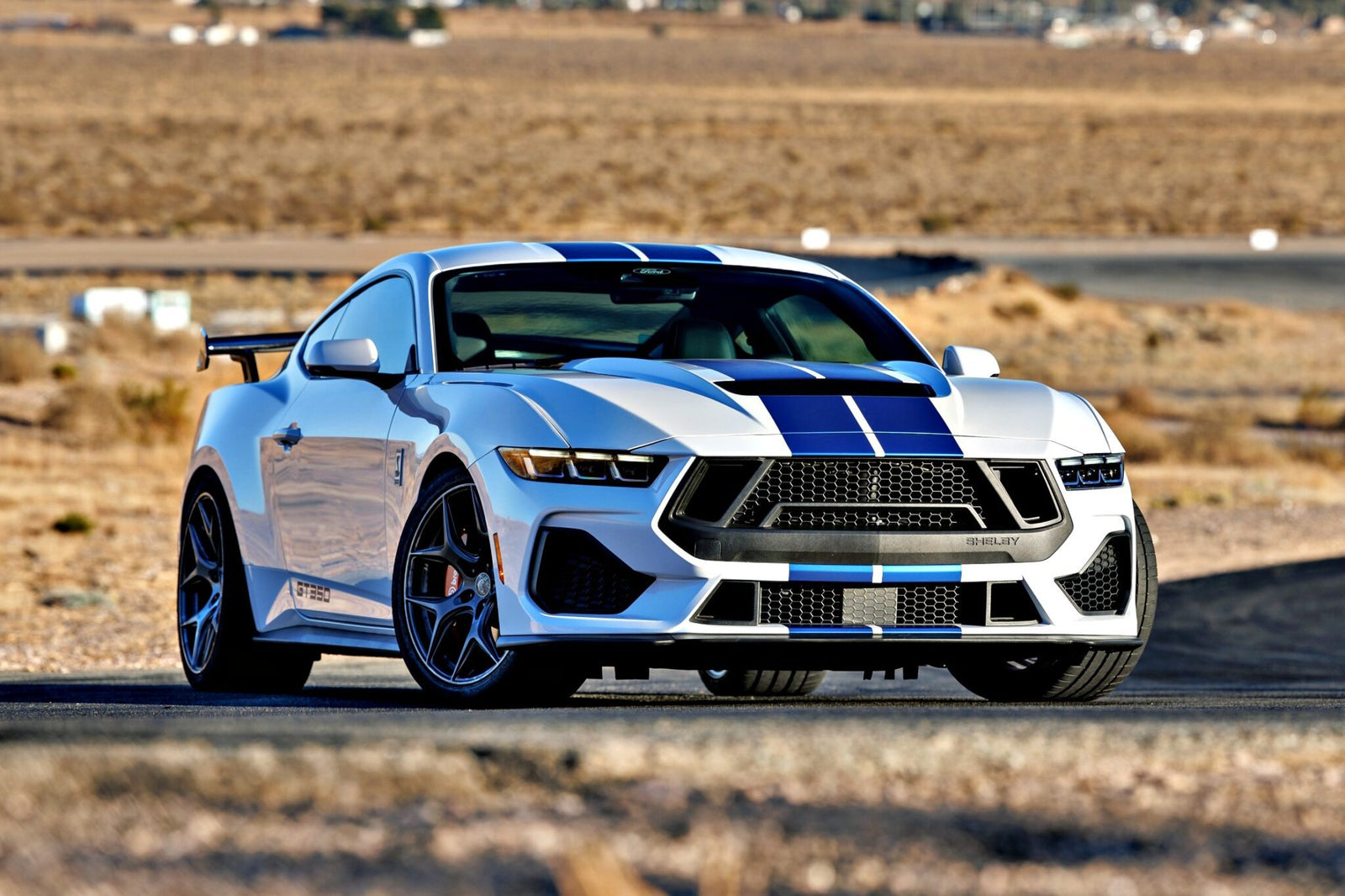 A white sports car with blue racing stripes, the Review: 2025 Shelby GT350, is parked on a paved road in a desert landscape with dry grass and distant buildings.