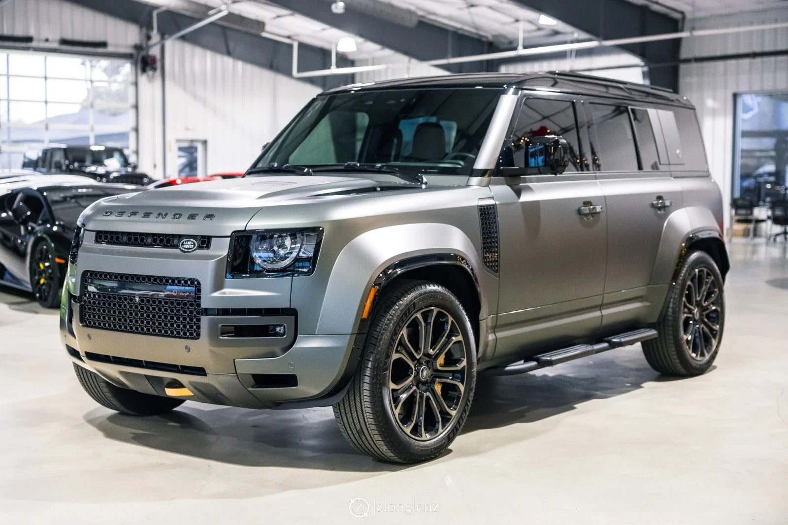 A silver Land Rover Defender SUV is parked indoors in a well-lit showroom, showcasing why it's considered among the best Land Rover vehicles, with other cars visible in the background.