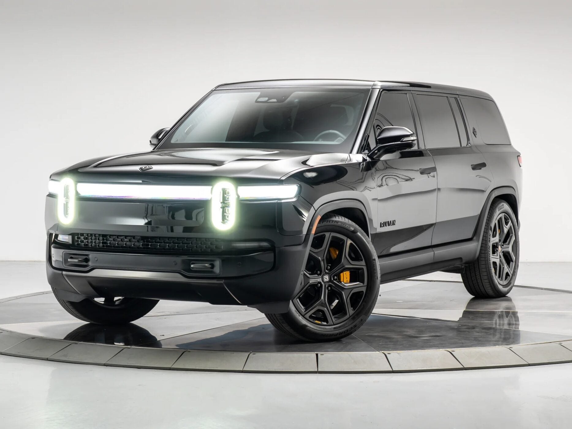 A black Rivian SUV, representing the new wave of affordable luxury SUVs, is parked on a circular platform in a studio setting, featuring distinctive illuminated front lights and black alloy wheels.