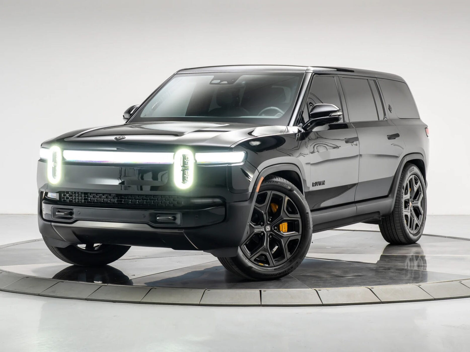 A black Rivian SUV, representing the new wave of affordable luxury SUVs, is parked on a circular platform in a studio setting, featuring distinctive illuminated front lights and black alloy wheels.