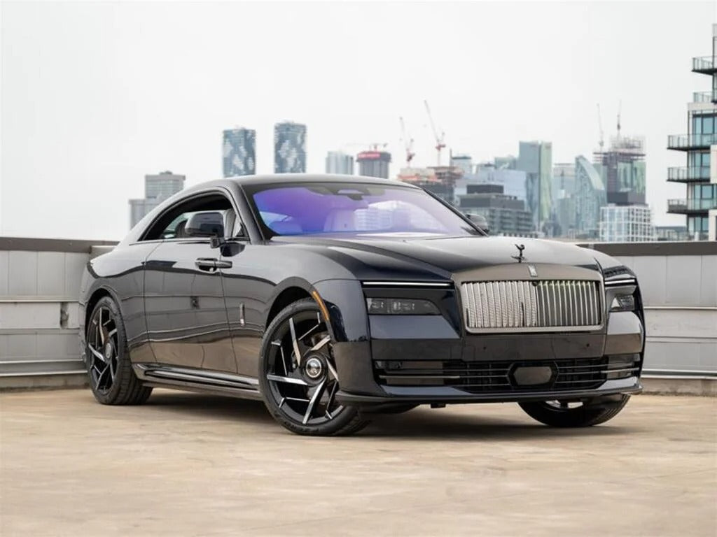 A black Rolls-Royce Spectre electric super coupe with a prominent grille is parked on a rooftop, set against a city skyline and construction cranes in the background.