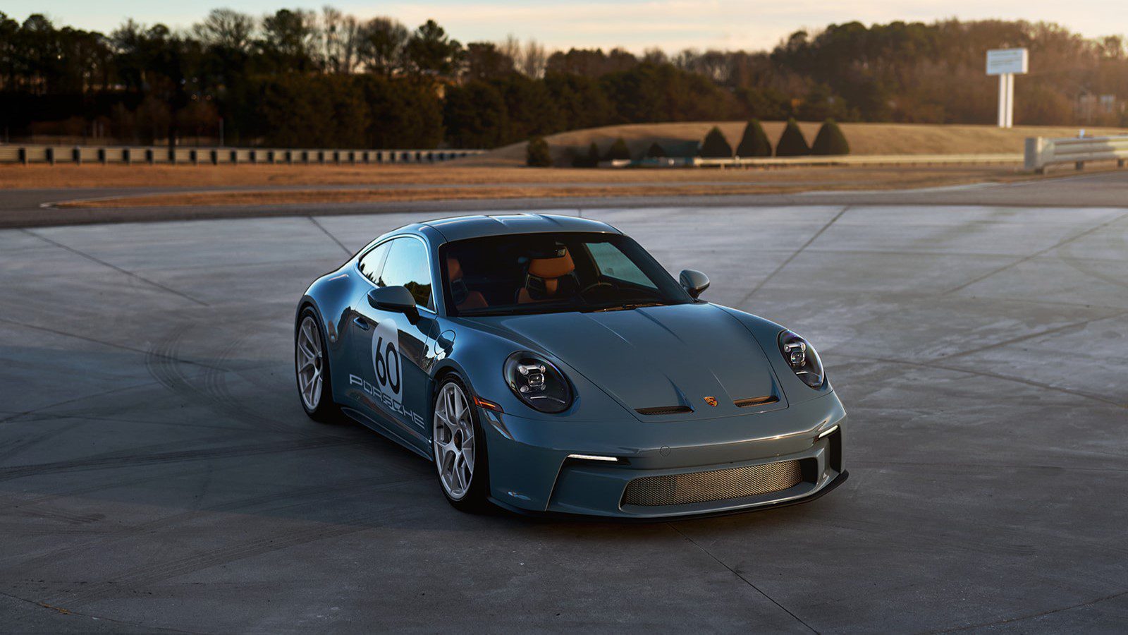 A gray Porsche 911 S/T sports car is parked on a large concrete surface, framed by lush trees and a clear sky in the background.