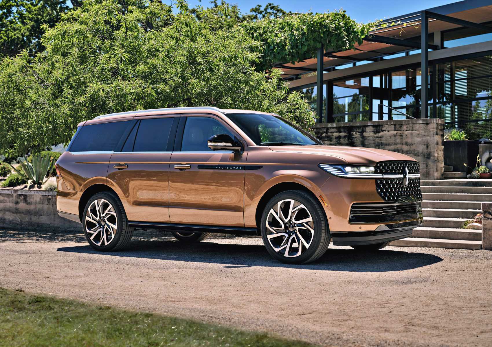 A bronze Lincoln Navigator SUV is parked on a gravel driveway in front of modern steps and greenery on a sunny day, echoing the comfort described in the Review: 2025 Lincoln Navigator L Black Label Coddles Like A Spa On Wheels.
