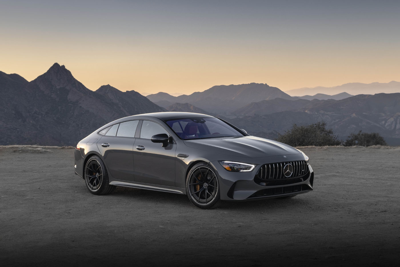 A sleek, gray Mercedes-AMG GT 63 S luxury sedan with E PERFORMANCE features is parked on a dirt road, framed by majestic mountains in the dusky backdrop.