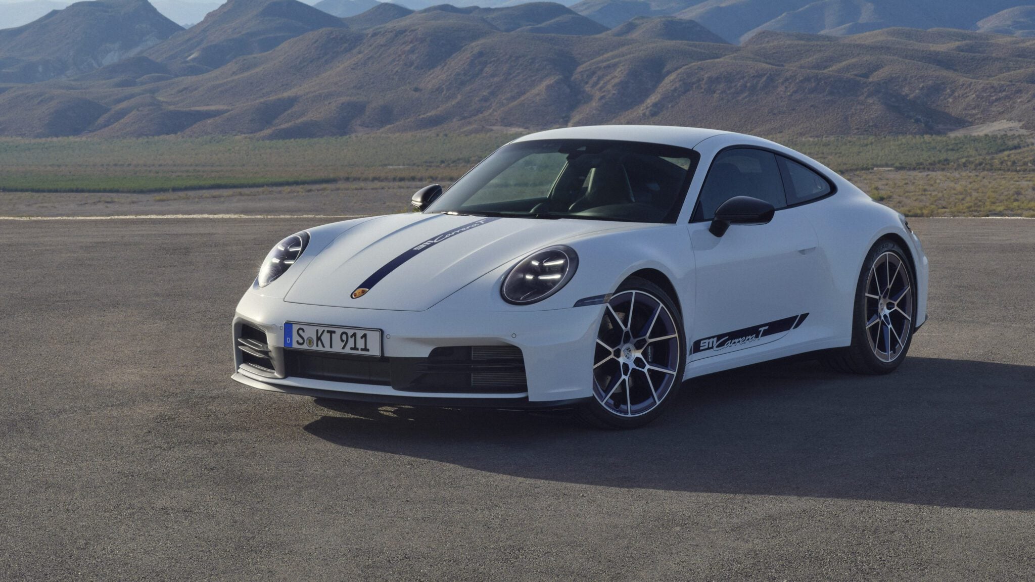 A white sports car is parked on a paved surface with mountainous terrain in the background.