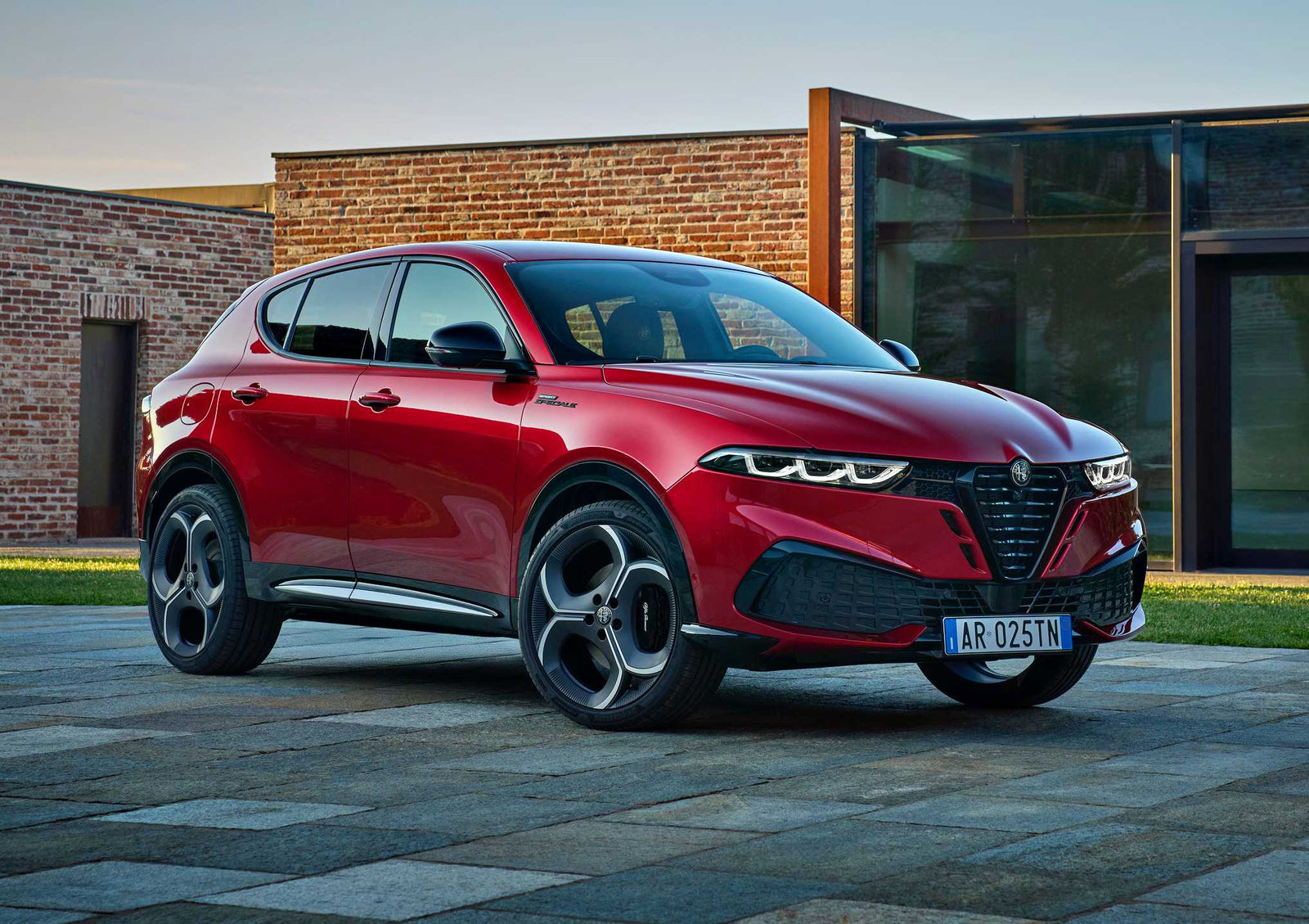 A red 2026 Alfa Romeo Tonale hybrid SUV is parked on a stone driveway in front of a modern brick and glass building.