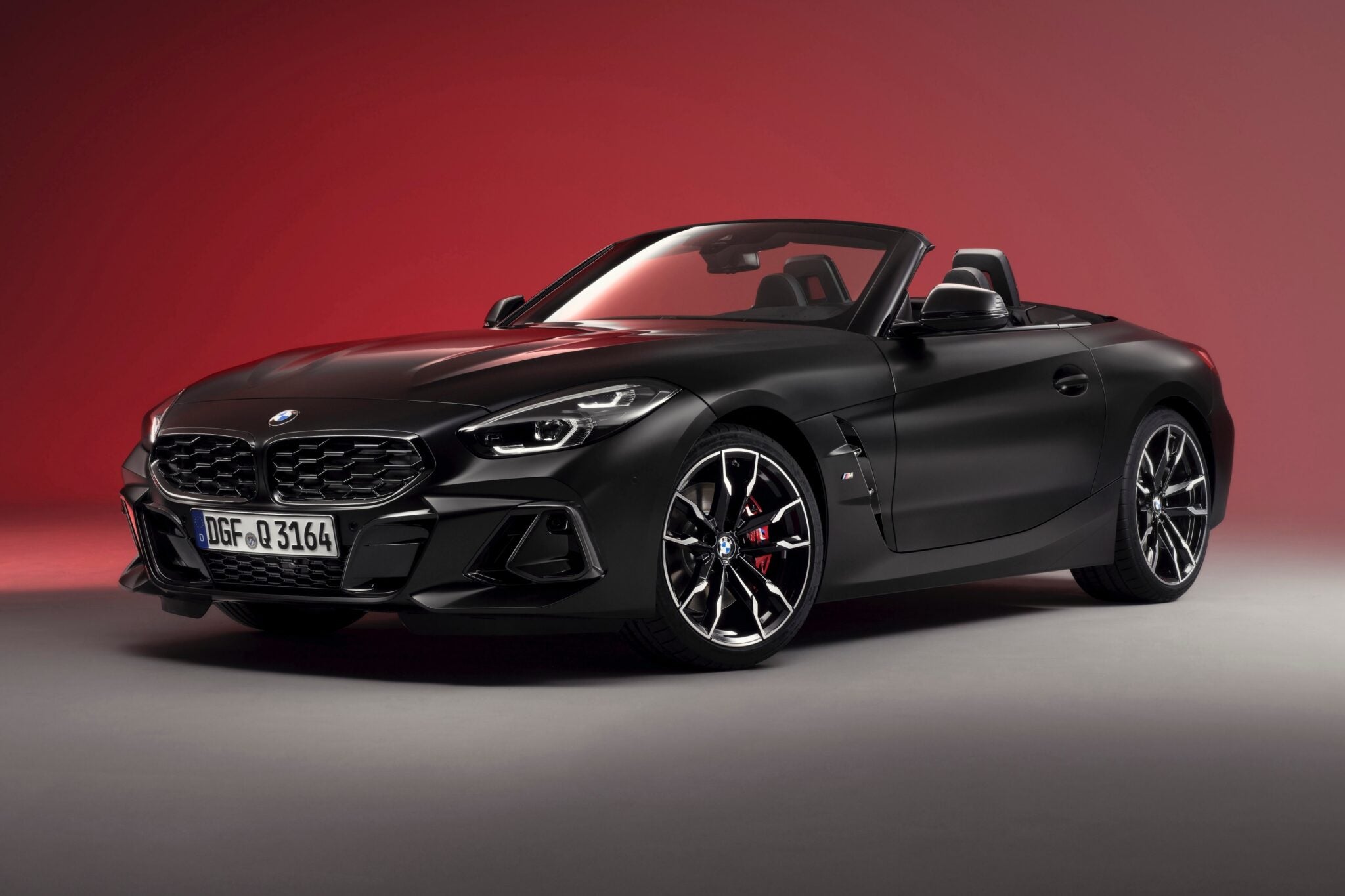A matte black BMW Z4 convertible sports car is shown in a studio with a red gradient background, highlighting the End of the Road: 2026 BMW Z4 Final Edition Breaks Cover in bold, striking style.