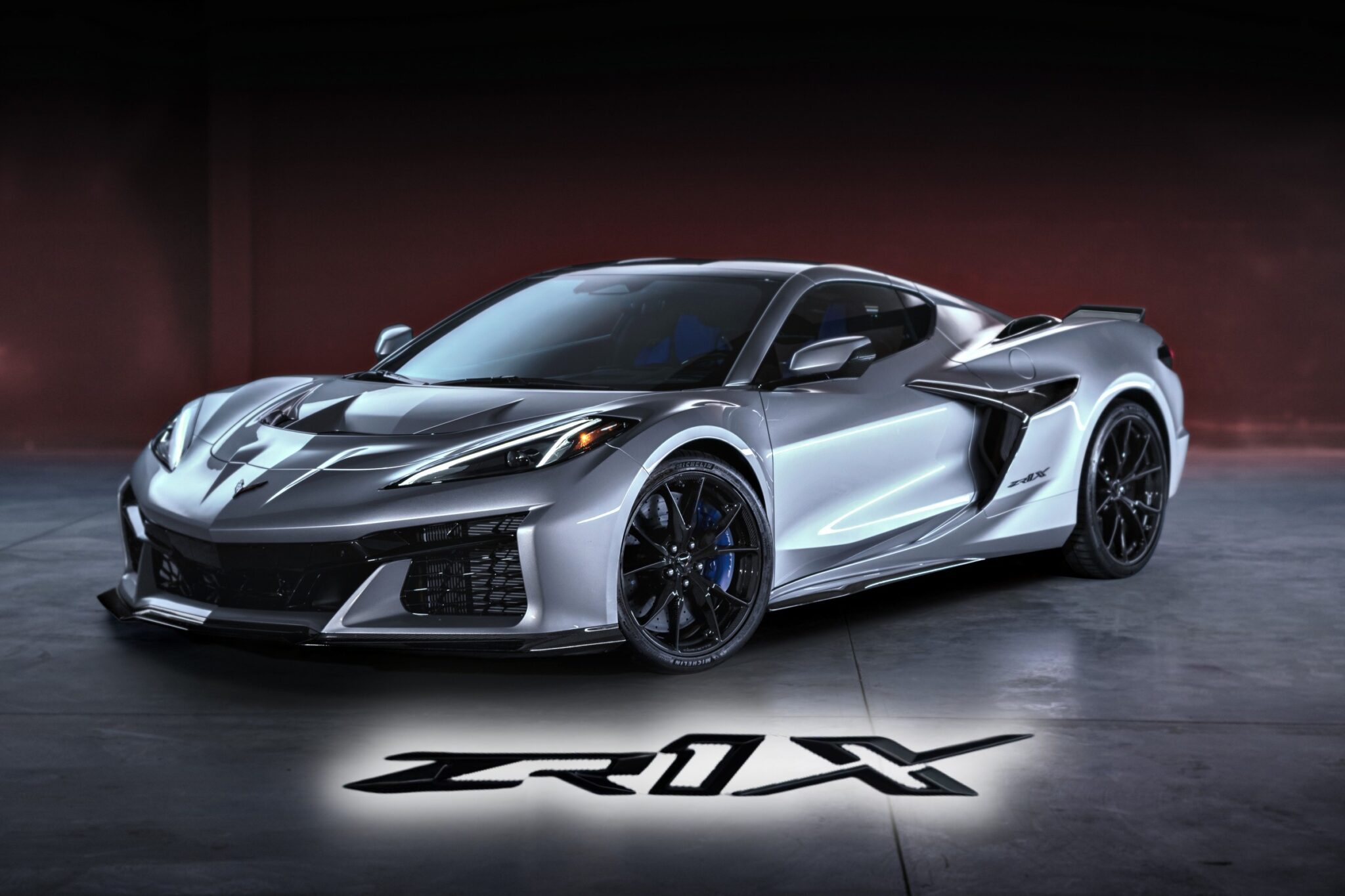A silver Chevrolet Corvette ZR1 sports car is parked indoors, its model name "ZR1X" illuminated on the floor in front. Revealed: 2026 Corvette ZR1X is the most powerful Chevrolet ever built.