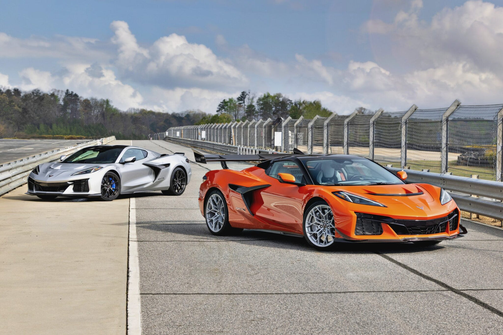 Two supercars—a silver and an orange Chevrolet Corvette ZR1X with 1,250-HP—are parked on a racetrack next to a fenced barrier under a partly cloudy sky.