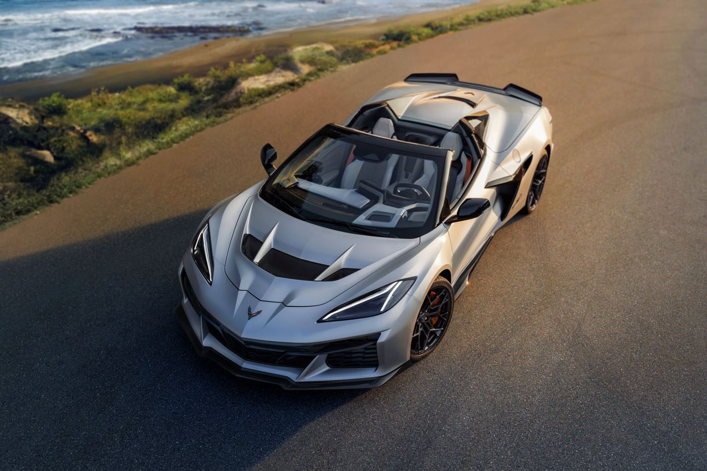 A Quail Silver Limited Edition Corvette ZR1X convertible is parked on a paved road near the coastline, with the ocean and greenery visible in the background, evoking the exclusive vibe of Monterey Car Week.