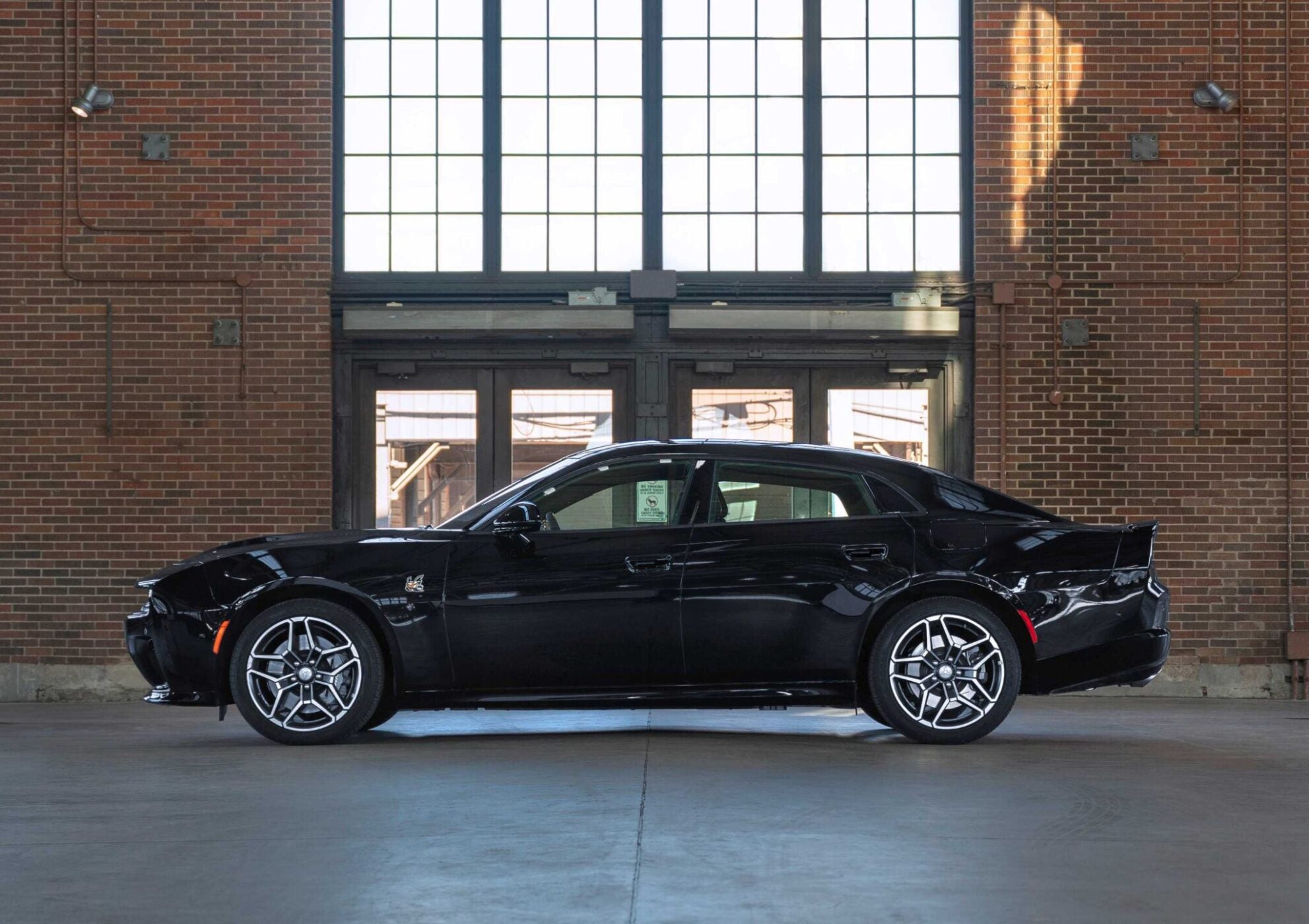 A powerful sedan in black is parked indoors, set against large windows and a brick wall.