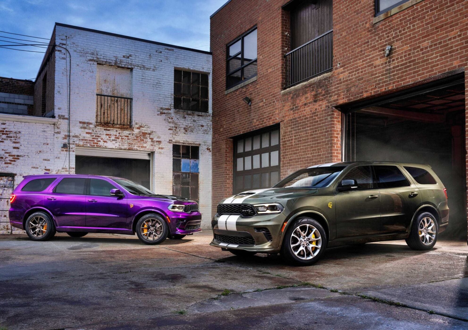 Two 2026 Dodge Durango Hellcat SUVs, one purple and one dark green with white stripes, are parked outside brick industrial buildings on a concrete lot, showcasing the bold presence of the Jailbreak edition.