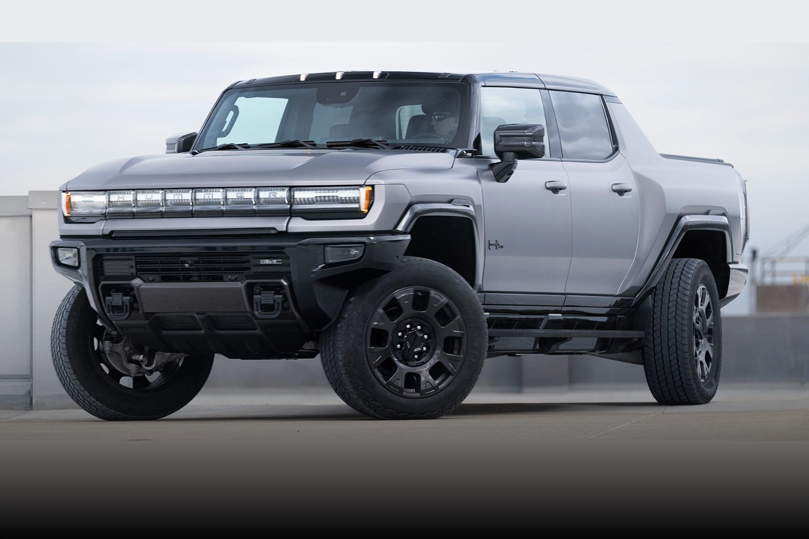 A silver GMC Hummer EV pickup truck, featured in the Review: 2026 GMC Hummer EV Pickup Opens A Toybox Of Excess, is parked outdoors on a concrete surface, viewed from the front left angle.