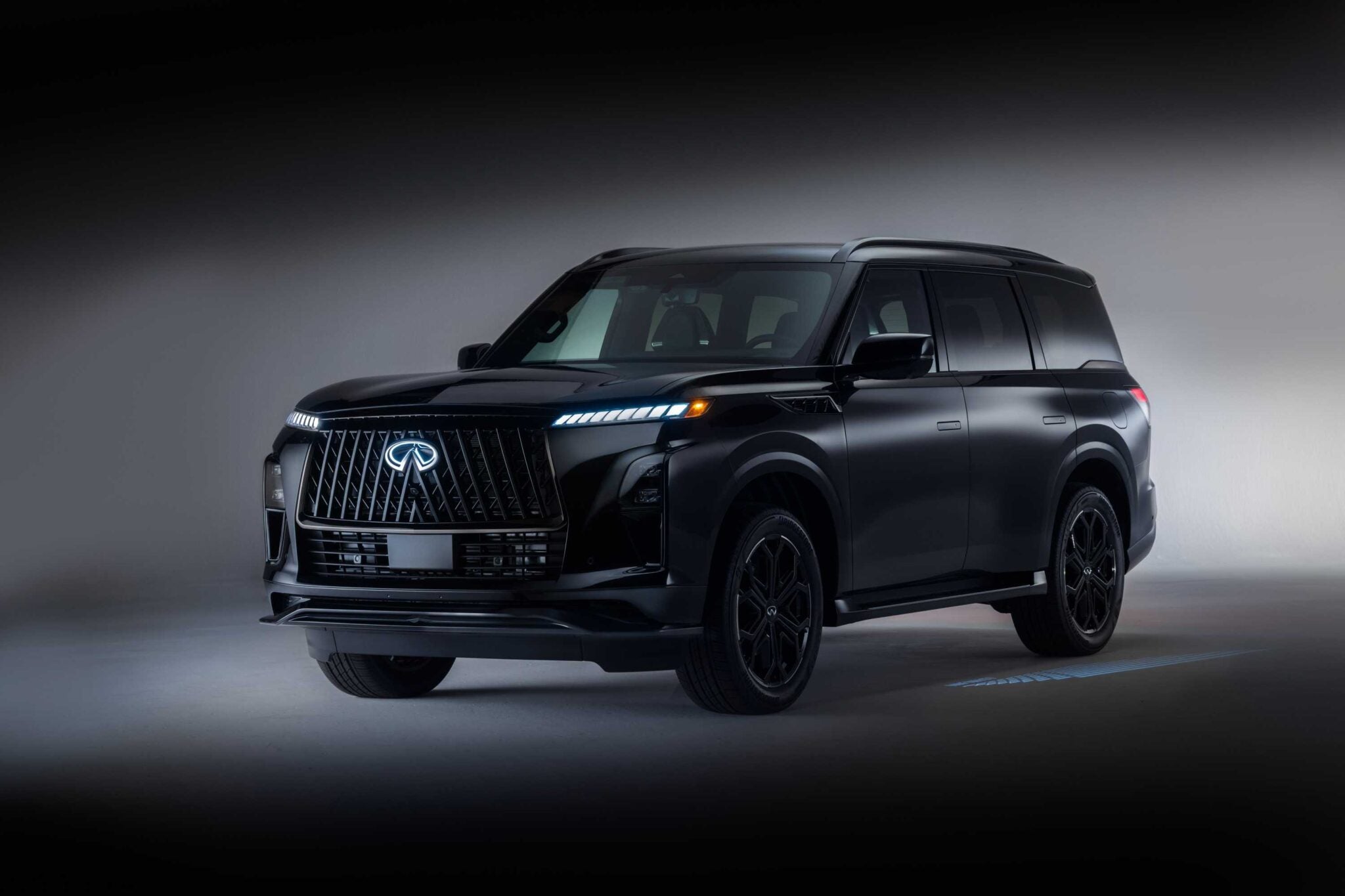 A black 2026 INFINITI QX80 SPORT Grade luxury SUV is parked in a studio setting, with soft lighting highlighting its front and side, truly upping the luxury game.
