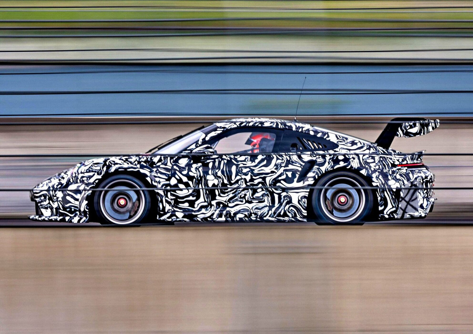 A next-gen race car with black-and-white camouflage wraps speeds along a track, motion blur highlighting its pace, and a large rear wing visible—hinting at innovations for the 2026 race season.