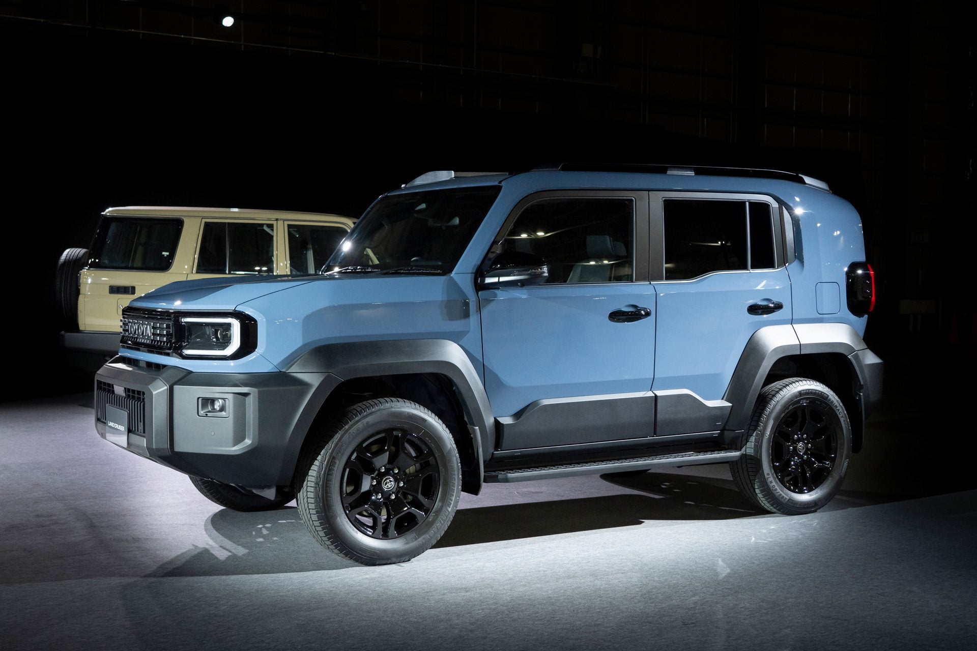 A blue 2026 Toyota Land Cruiser FJ compact SUV is displayed indoors under bright lighting, projecting a capable and rugged stance, with a beige SUV visible in the background.