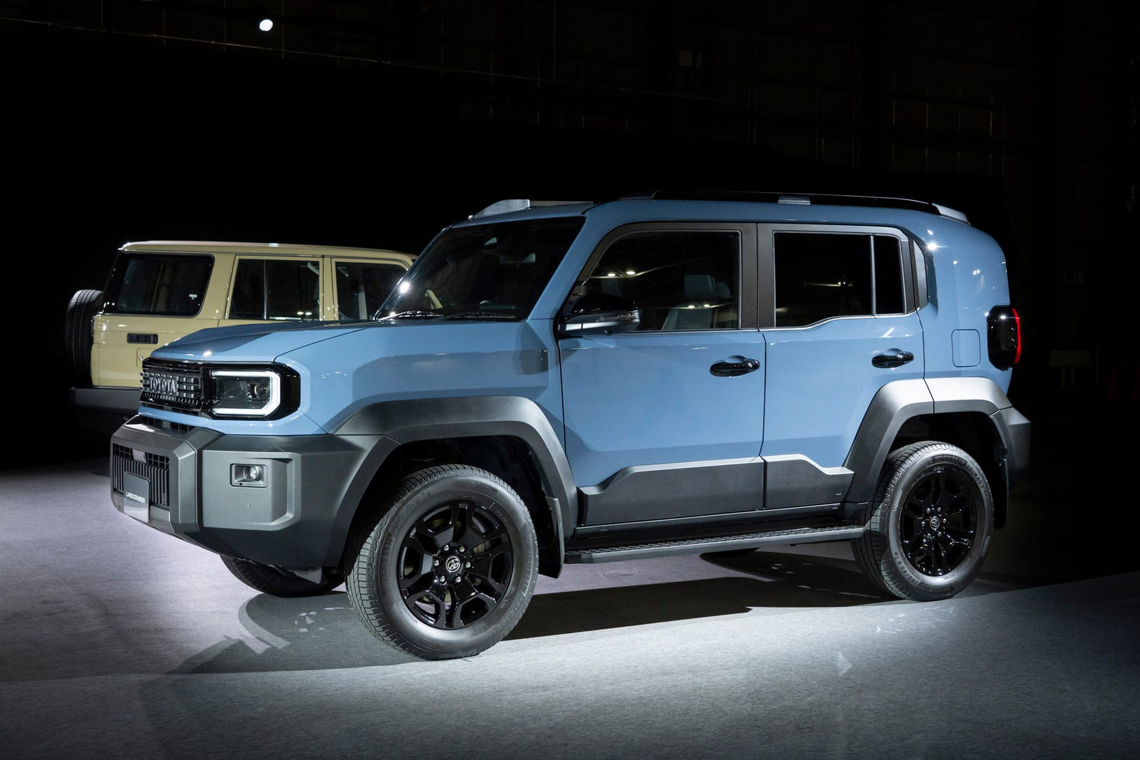 A blue 2026 Toyota Land Cruiser FJ compact SUV is displayed indoors under bright lighting, projecting a capable and rugged stance, with a beige SUV visible in the background.