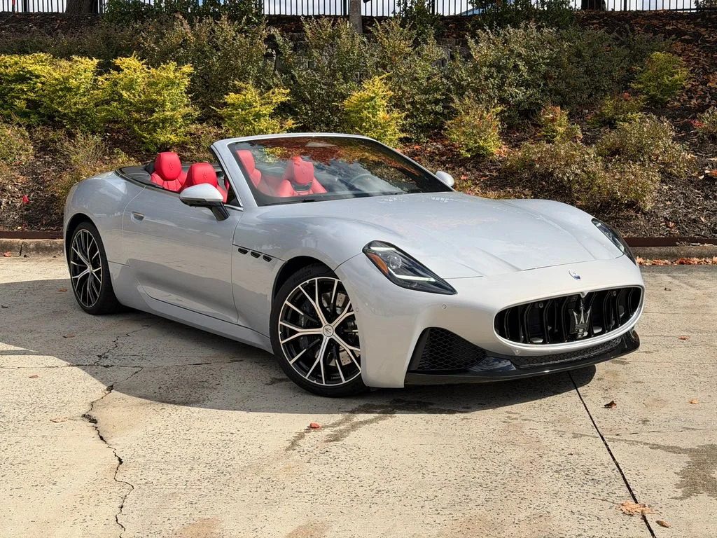 A silver Maserati convertible sports car with a red interior is parked on a concrete surface near bushes and a fence, showcasing true Italian luxury.