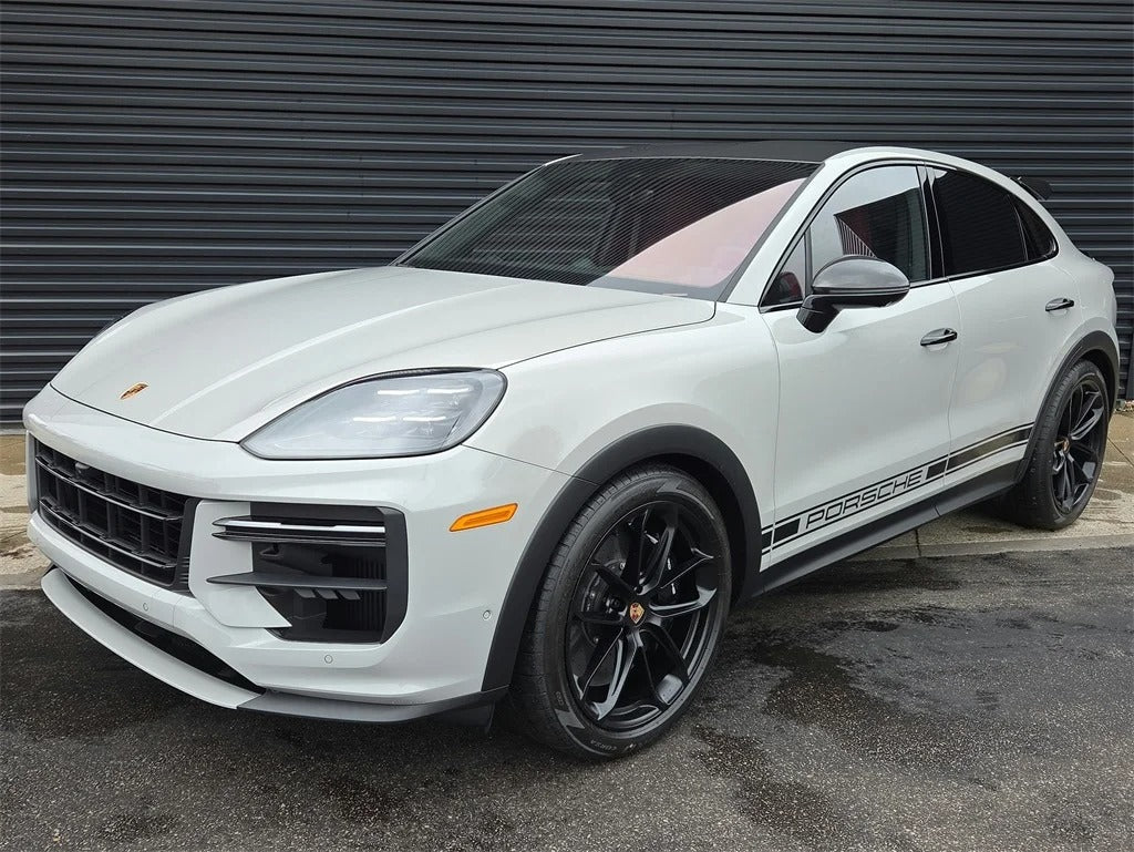 A white Porsche Cayenne Turbo GT, a true luxury performance SUV with black wheels and "Porsche" side decals, is parked on wet pavement in front of a dark corrugated metal wall.