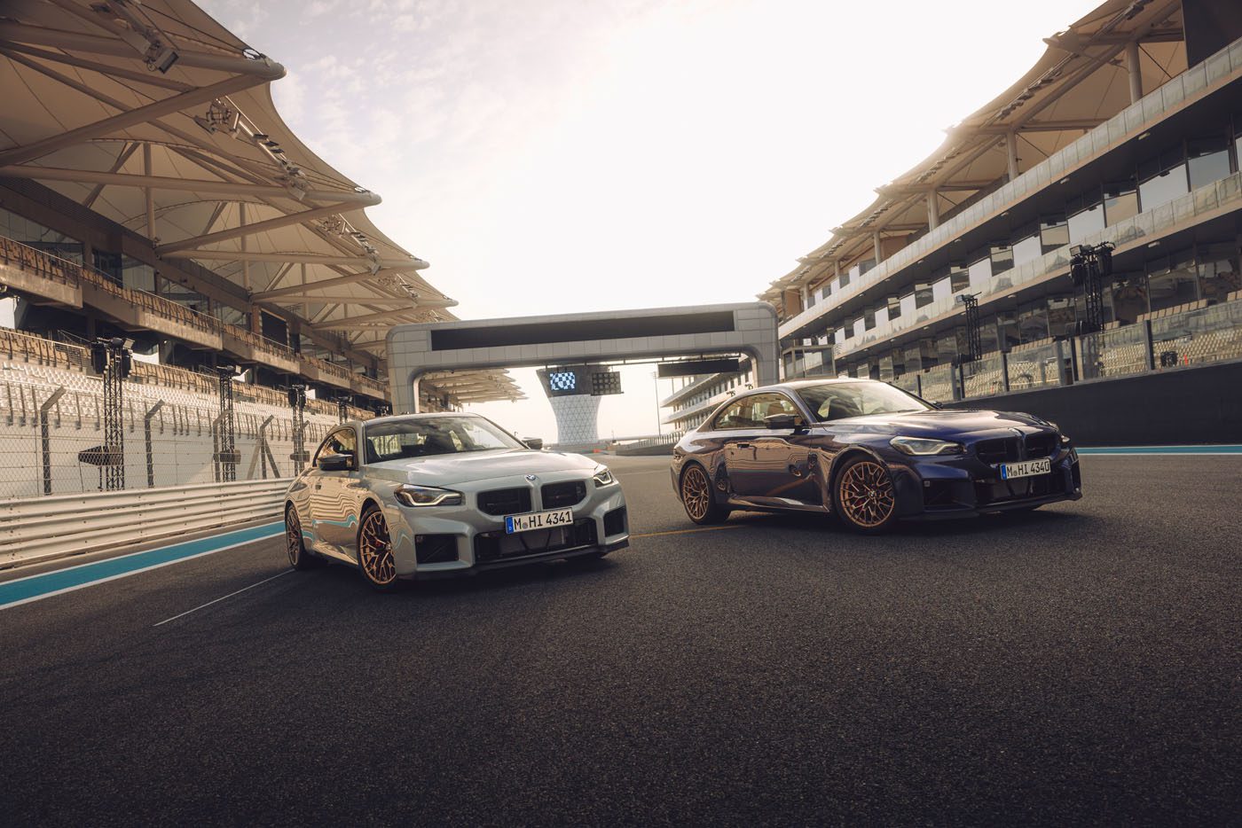 Two BMW cars are parked side by side on a racetrack at Monterey Car Week 2025, with empty grandstands and a modern structure in the background under a bright sky.