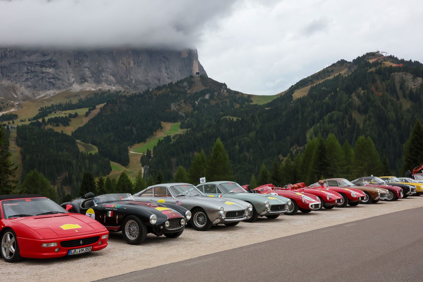 A row of classic and vintage sports cars, part of the Ferrari Cavallcade, parked by a mountain road in South Tyrol, Italy, with forested hills and cloudy skies in the background.