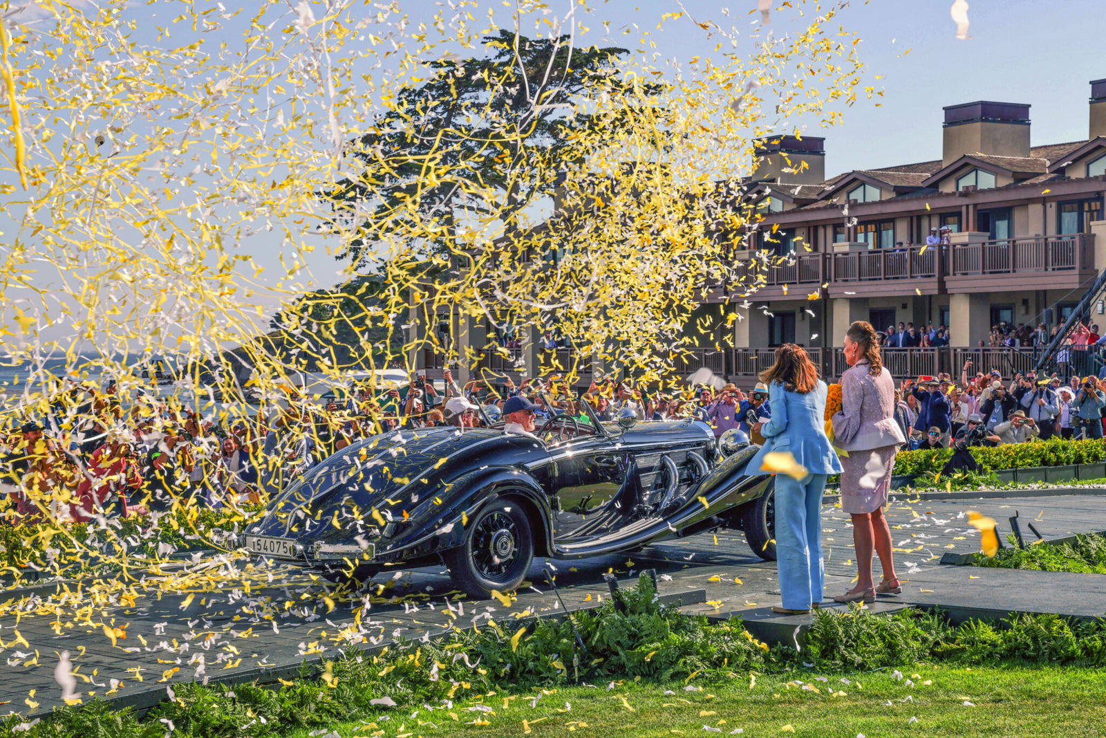 A vintage car with a glossy black finish is celebrated onstage at the illustrious Concours d'Elegance, as yellow confetti falls. A crowd is gathered nearby, with buildings in the background, embodying the essence of Pebble Beach's upper echelon.