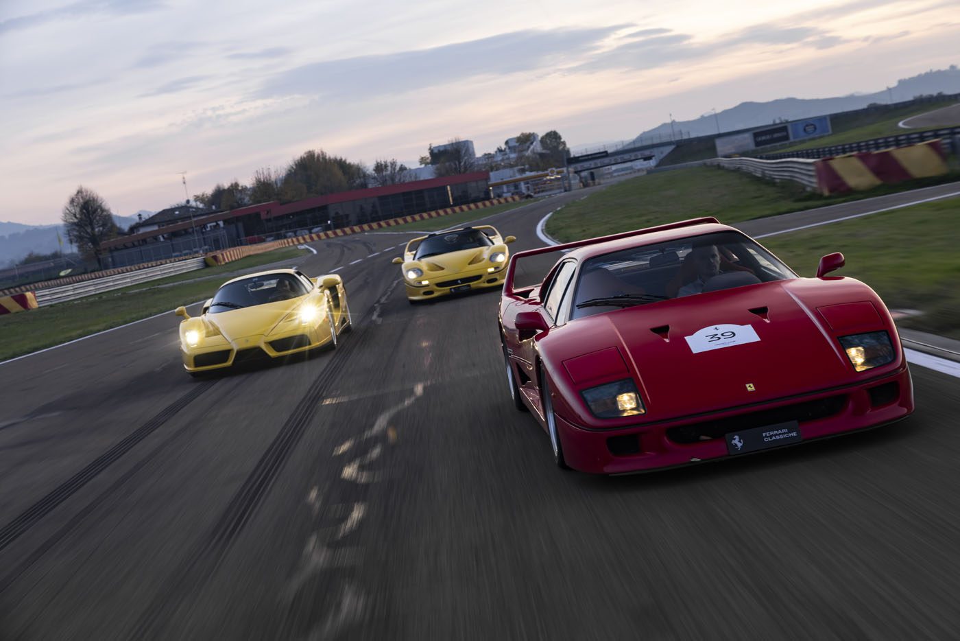 Three sports cars, two yellow and one red, drive on a racetrack at sunset. Grass and buildings are visible in the background.