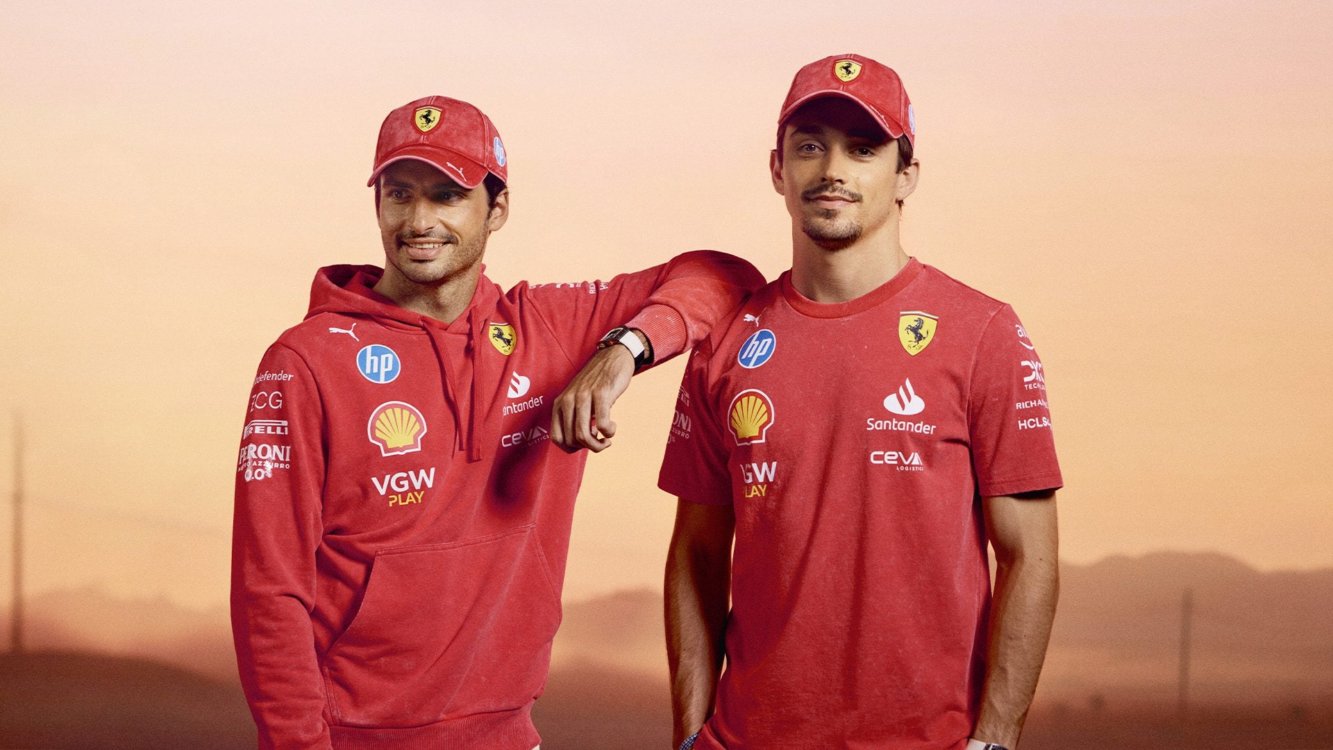 Two individuals in red Scuderia Ferrari team uniforms and caps stand against a sunset background, embodying the spirit of the Desert Sun Collection.