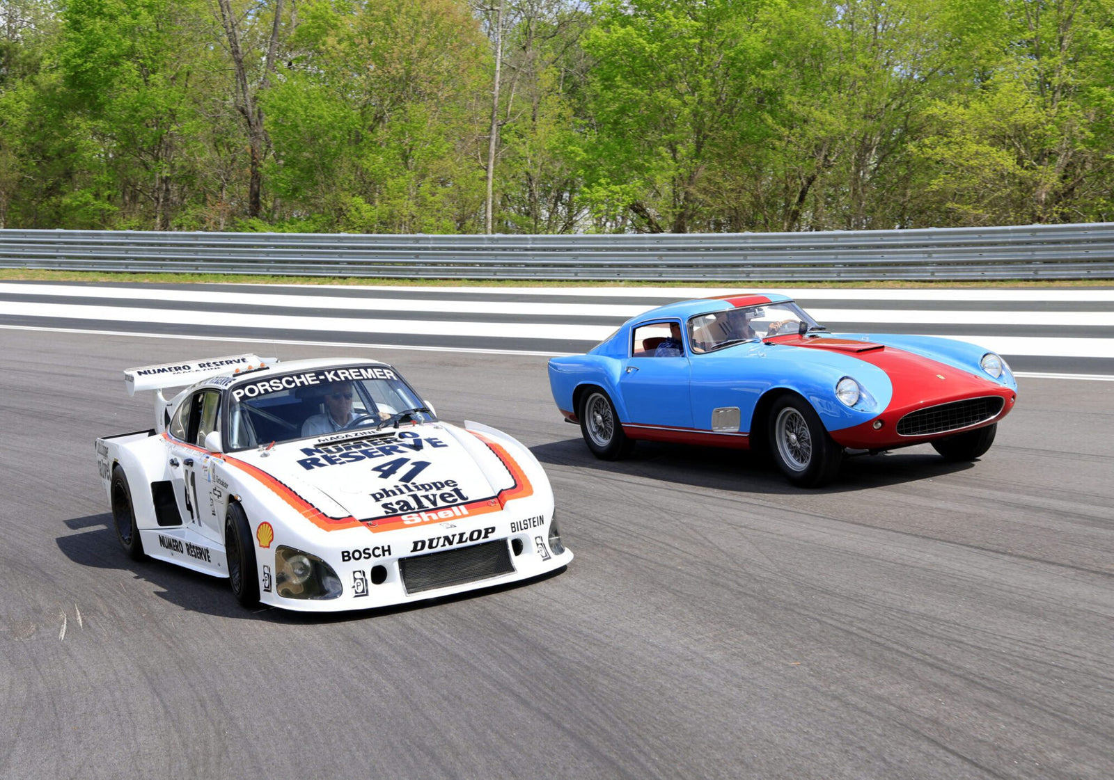 A white Porsche race car and a blue classic Ferrari sports car drive side by side on a track with greenery in the background.