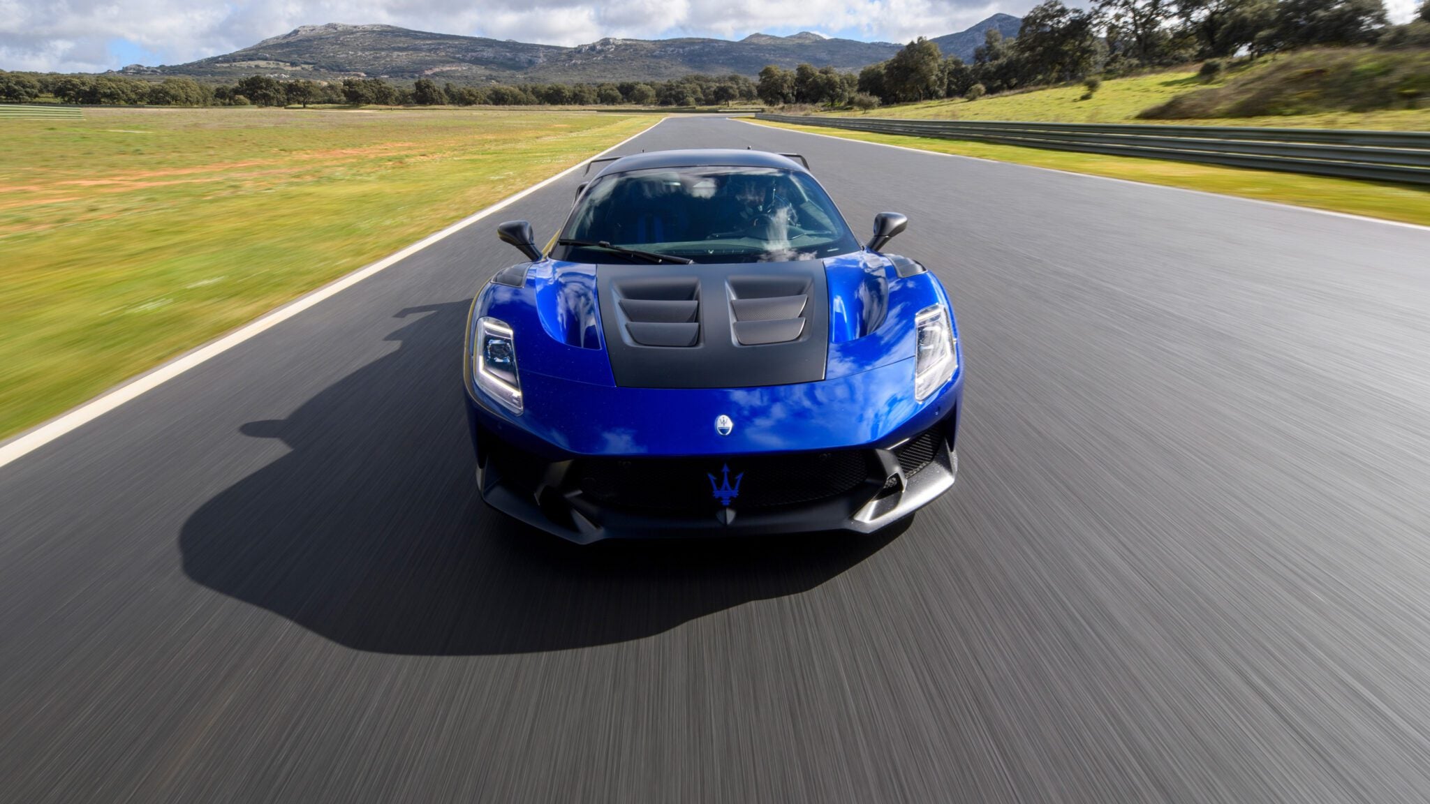 A blue sports car drives on a race track with mountains in the background under a partly cloudy sky.