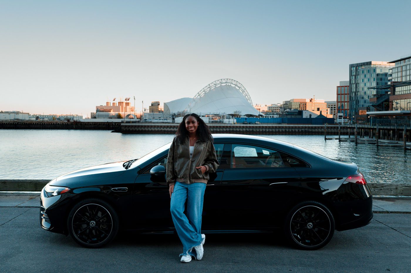 A woman, possibly a Global Brand Ambassador, stands in front of a parked black Mercedes-Benz sedan by the waterfront, with modern buildings and a distinctive arched structure in the background.