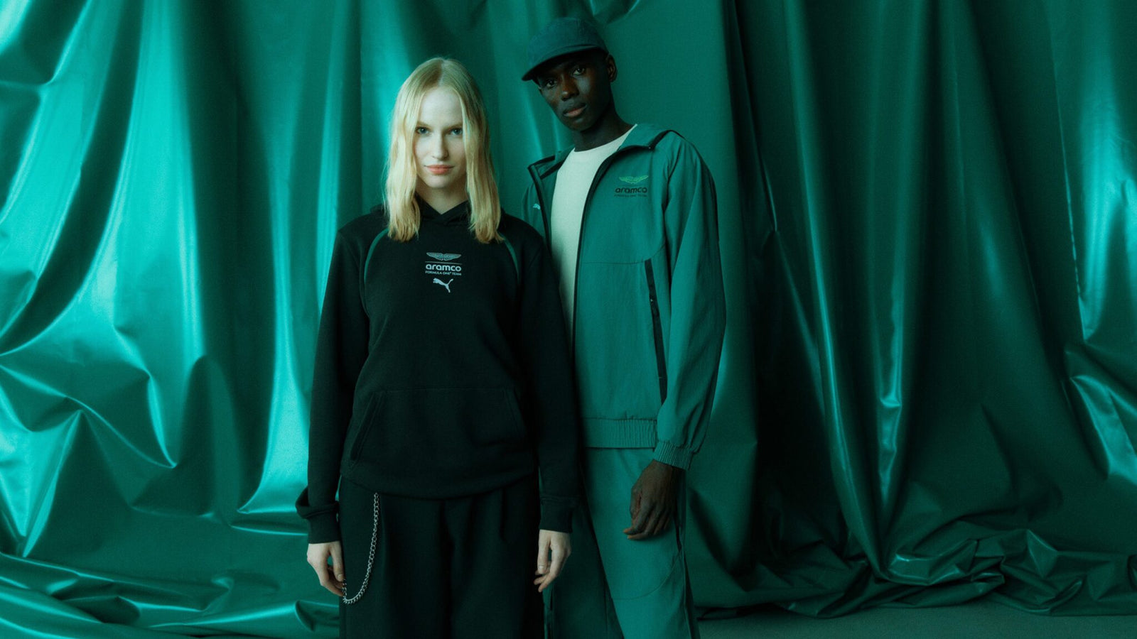 Two people stand in front of a green draped background; one wears a black hoodie and skirt, while the other sports a green jacket, pants, and cap from the PUMA Motorsport F1 Team Collection.