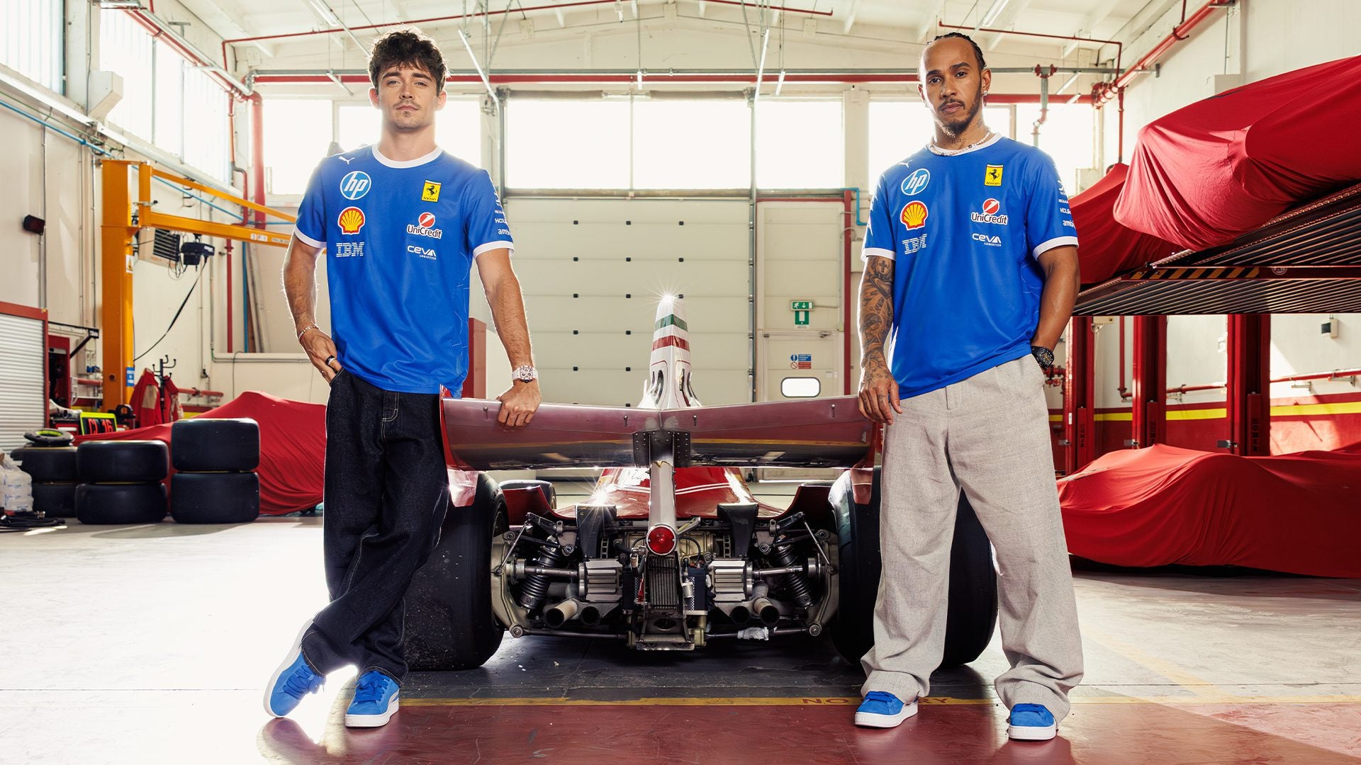 Two men in blue PUMA x Ferrari shirts stand on either side of a Formula 1 car in a garage, with covered vehicles and tires visible in the background.